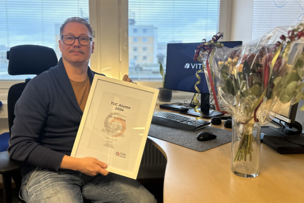 Dan Stjärnborg sitter på sitt kontor och håller upp diplomet för utmärkelsen TUC Alumn. I förgrunden syns en stor bukett och bakgrunden hans dator och ett fönster.