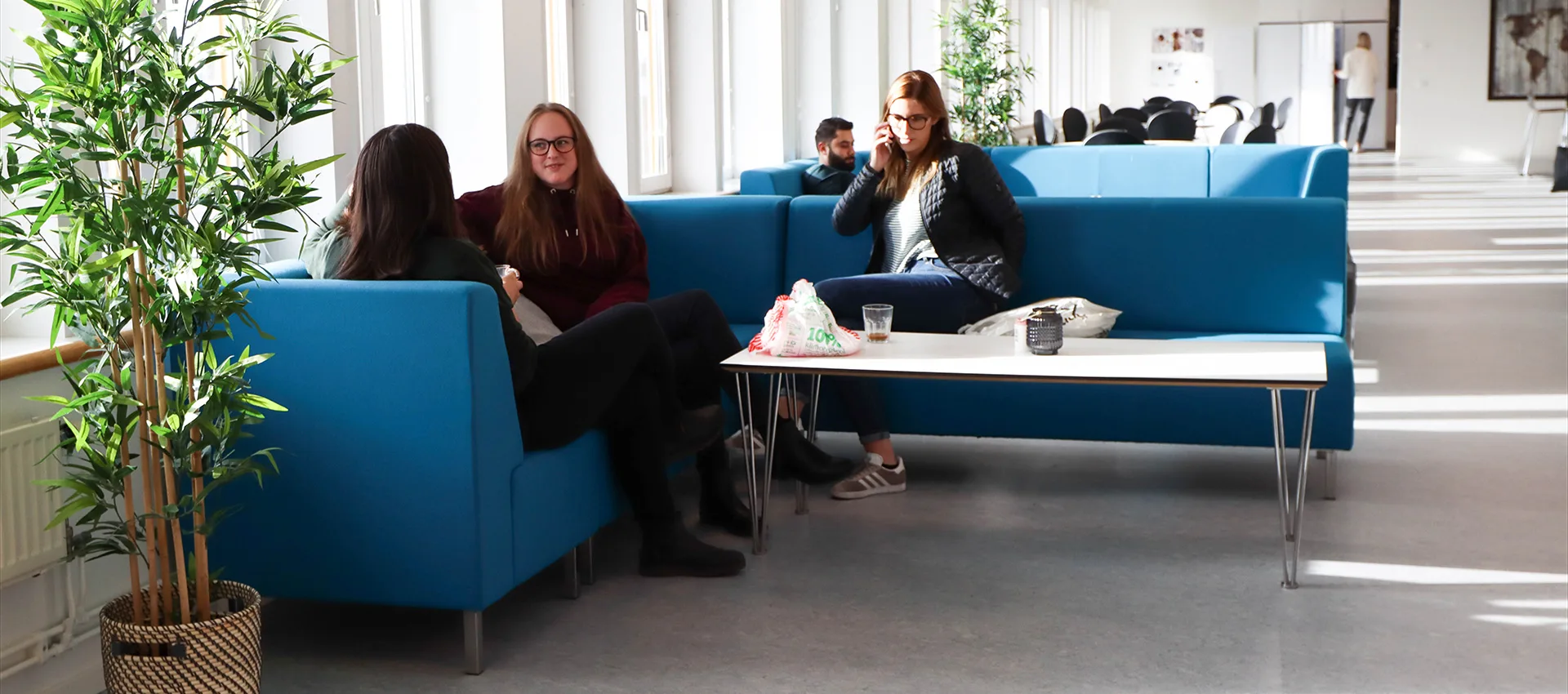 Två kvinnliga studerande sitter i en blå soffa och pratar med varandra. En tredje kvinna syns också, hon pratar i telefon.